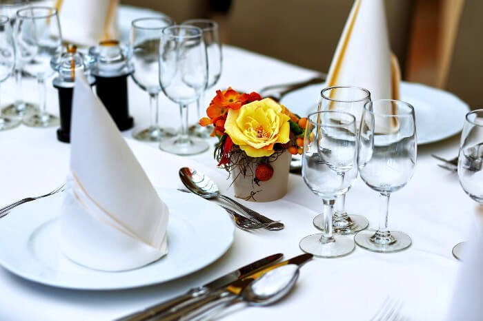 A formal table setting with a white tablecloth, folded white napkins on white plates, silver cutlery neatly arranged, multiple glasses for beverages, and a small floral centerpiece—suggesting a refined setting for a wedding or banquet